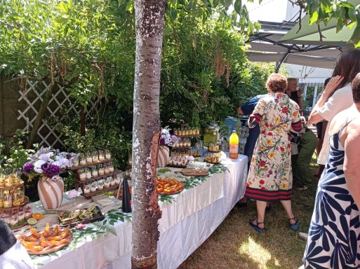 Buffet extérieur coloré avec plats variés, fleurs et convives en arrière-plan.